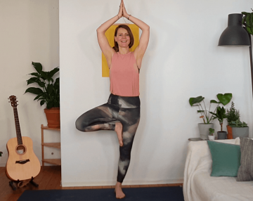 Person in Yogakleidung in Pose des Baumes vor weißer Wand, rechts daneben Sofa mit Kissen sowie Pflanzen, links davon Gitarre
